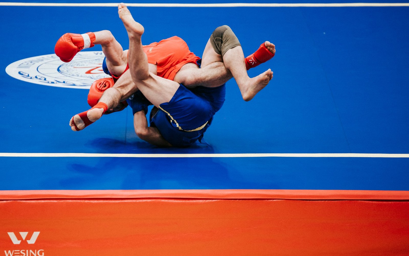 Two grapplers wearing red gloves tangle mid-throw on a blue competition mat