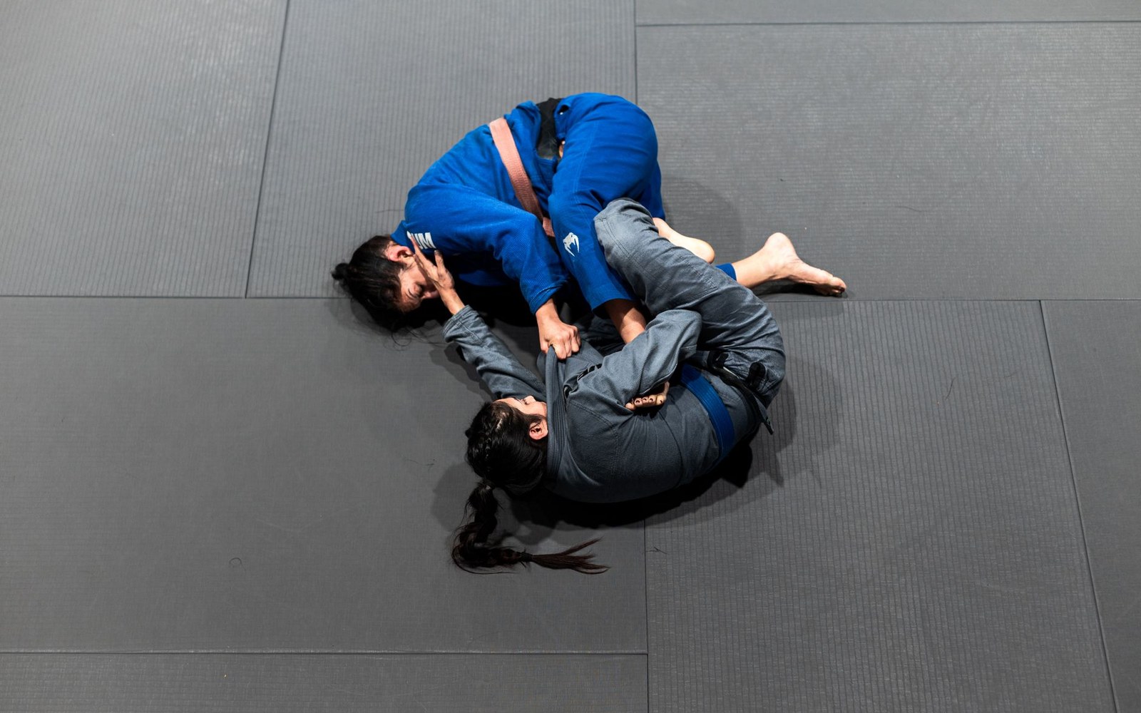 Two grapplers in blue and grey gis practice a ground submission hold on a mat