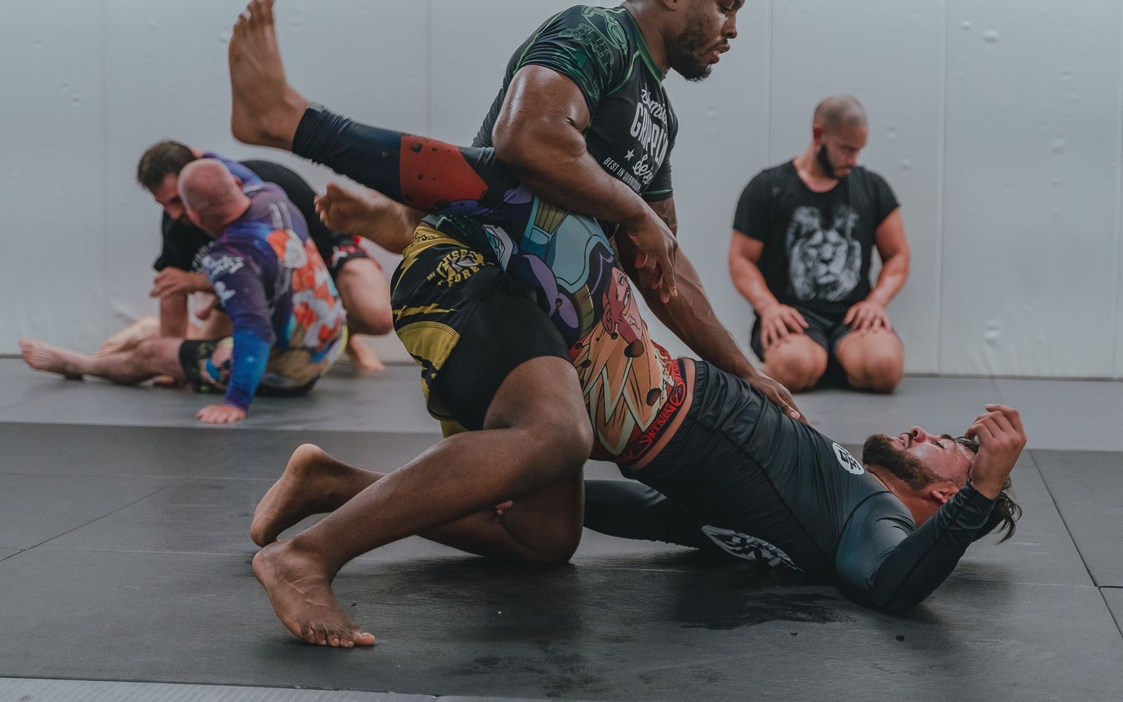 Grapplers practice no-gi jiu-jitsu submissions on mats while others train in the background