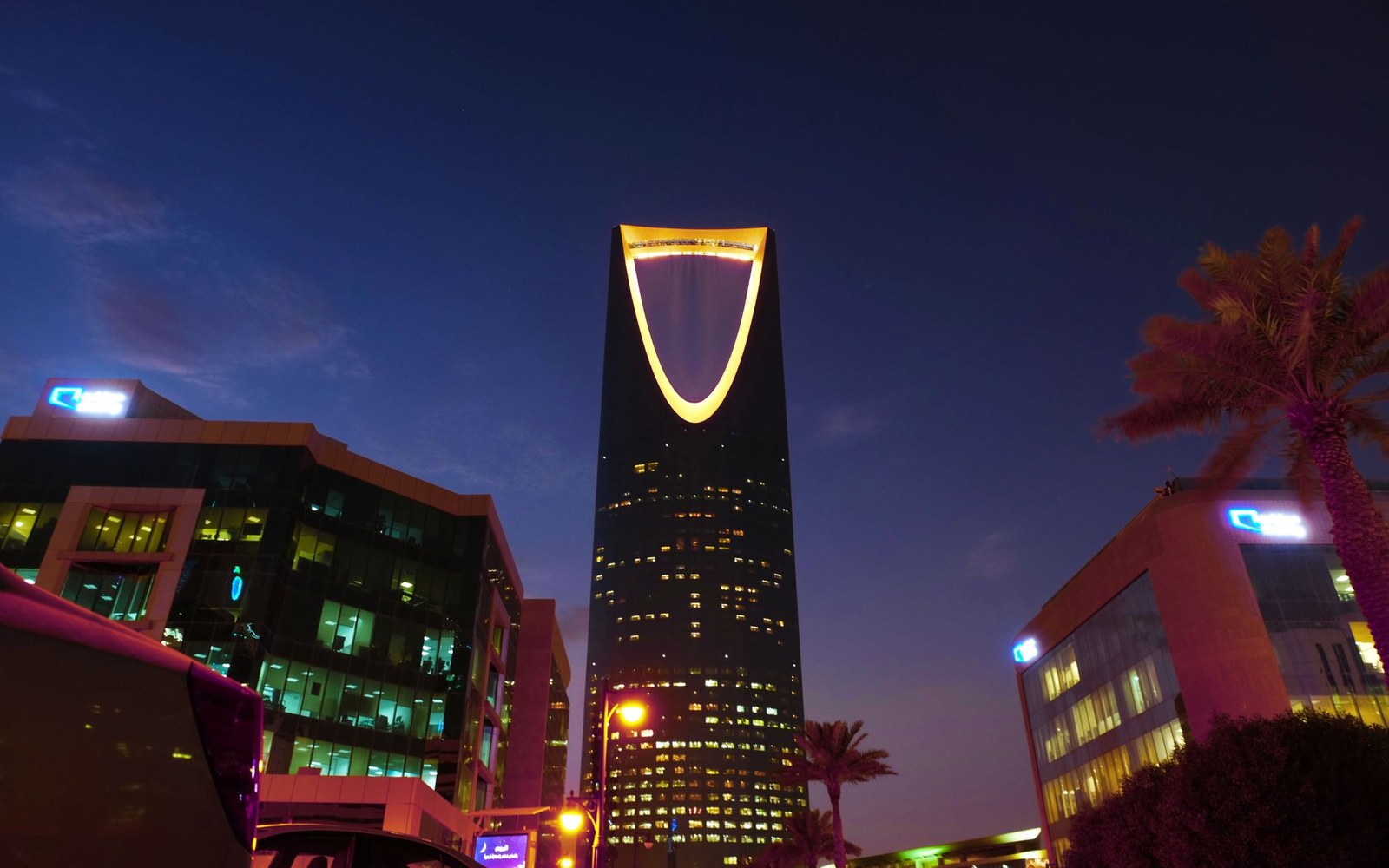 Kingdom Tower's illuminated arch rises above lit office buildings at dusk in Riyadh