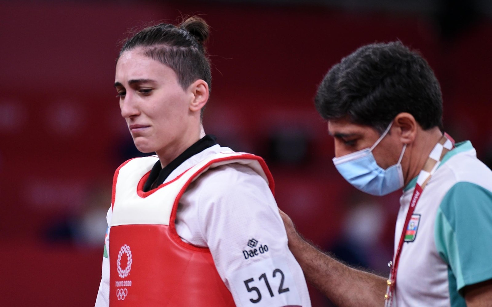A tearful taekwondo athlete in a red scoring vest walks off as a coach touches her shoulder