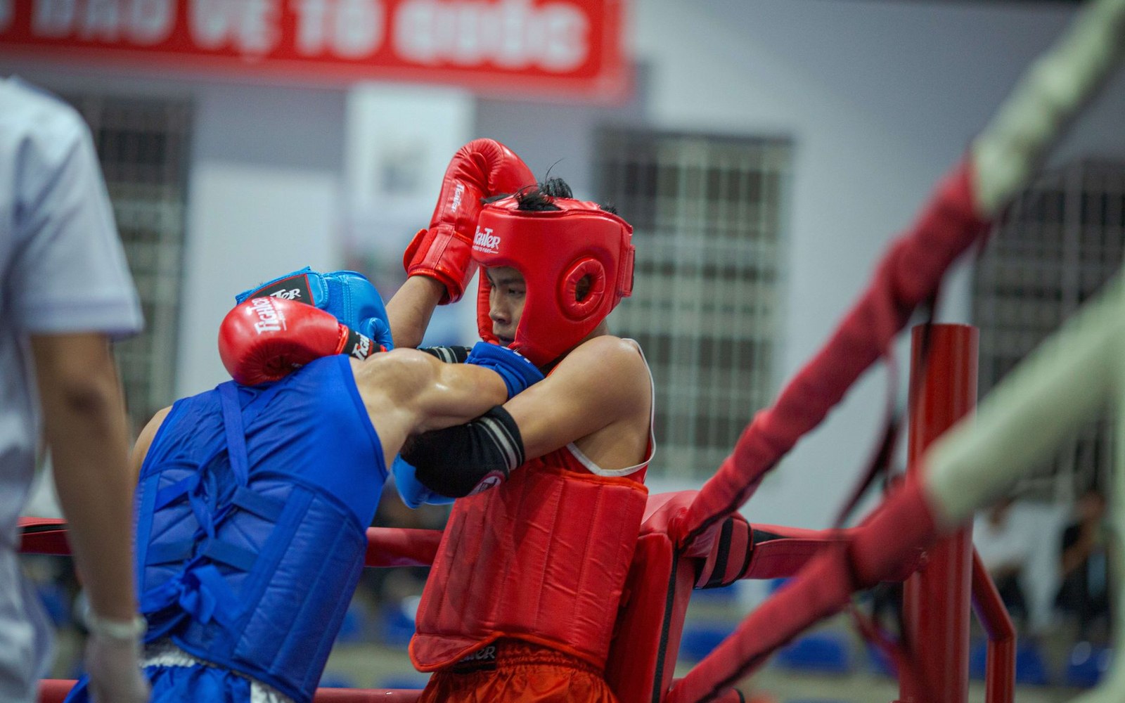 Two amateur boxers in red and blue headgear clinch against the ring ropes